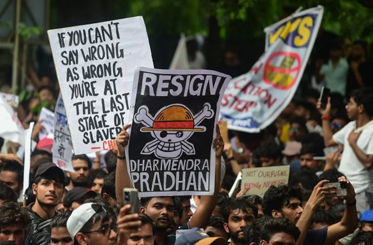 A protest crowd holding hand-drawn placards; the largest reads 'Resign Dharmendra Pradhan', another reads 'If you can't say wrong as wrong you're at the last stage of slavery'.