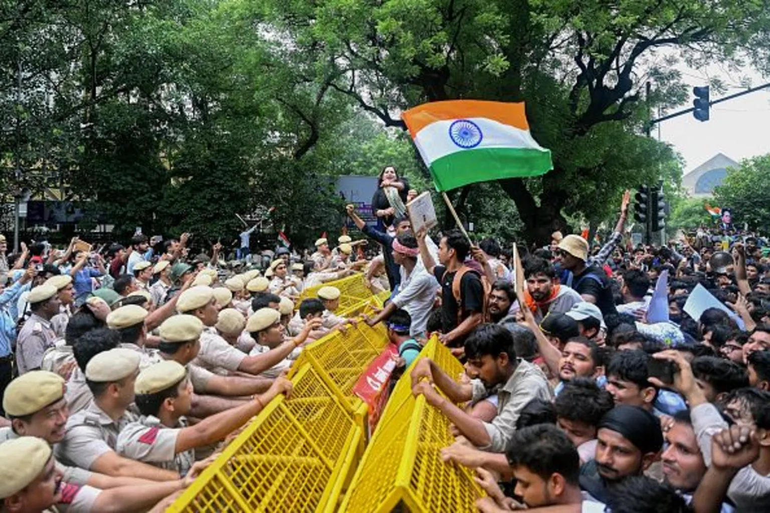 Students pressing against yellow police barricades at a protest in New Delhi, one waving the Indian tricolour; rows of police push back from the other side.